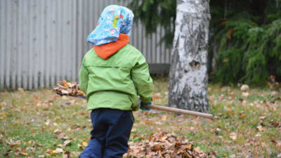 Ett barn med grön jacka och huckle fotat bakifrån. Barnet bär en kratta och i bildens bakgrund finns en löv. 