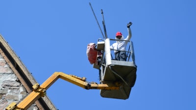 En man iklädd heltäckande kläder står i en skylift bredvid ett tak och vinkar mot kameran.