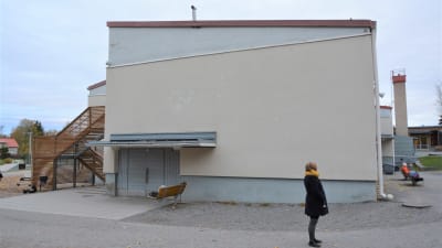Ändan av en stor skolbyggnad. Framför betongväggen står en kvinna och ser mot sidan.
