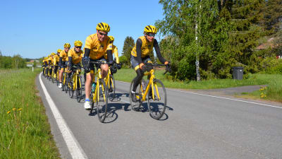 Ett gäng cyklister i gula skjortor och gula hjälmar cyklar på en landsväg i ett grönt Sommarfinland.