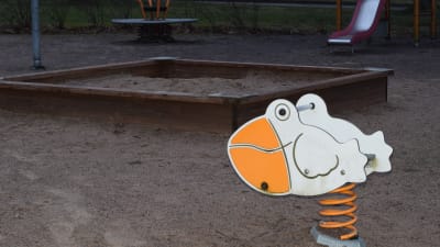 En gunga för barn som föreställer en anka eller pelikan i en lekpark. Sandlåda och rutschbana i bakgrunden. Inga barn.
