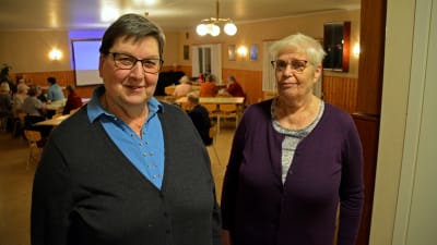 Två kvinnor står i dörröppningen till en stor sal där människor sitter runt bord.