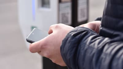 Händer håller i en smarttelefon vid en laddstation för elbilar.