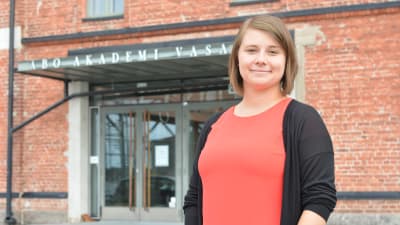 matilda ståhl bär en röd tröja och står utanför åbo akademis tegelbyggnad i vasa. i bakgrunden ser man ingången och ovanför den står det åbo akademi vasa
