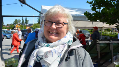 titti edfelt har vitt hår, vit halsduk, grå jacka och ler mot kameran. I bakgrunden syns andra människor i festivalvimlet.