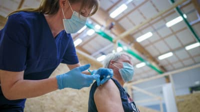 En man blir stucken med en spruta innehållande coronavaccin, av en sköterska i mörkblå kläder och ljusblå gummihandskar.