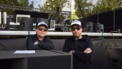 Två personer med solglasögon står framför en scen.