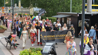 Färggrant klädda personer går i en prideparad, en del håller i en svart stor banderoll med texten Åbo Akademis studentkår