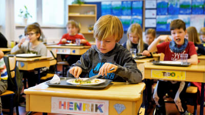 Många elever äter lunch vid sina pulpeter.