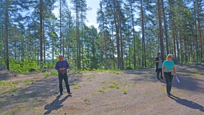 Två kvinnor och en man går omkring på ett område med sand omgivna av en tallskog.