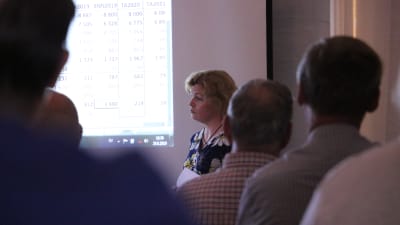 En kvinna med trumpen står framför en videoduk och presenterar dystra ekonomiska siffror.