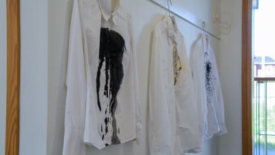 Tre vita kragskjortor hänger på en vit vägg. En av skjortorna har svart färg spilt över sig, en har ett hål bränt vid bröstet och en har svarta små bollar sydda på sig. 