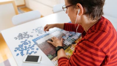 Keski-ikäinen nainen kuuntelee audiosisältöjä Yle Areenasta samalla kun kasaa palapeliä pöydän ääressä.