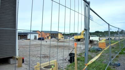 Byggarbetsplats på grusplan bakom stängsel. Bostadshus i bakgrunden.
