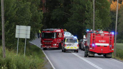 Två stora röda brandbilar och en huvudsakligen vit polisbil på en smal väg omgiven av träd och en åker.  Brandkåren och polisen på olycksplatsen i Borgå.