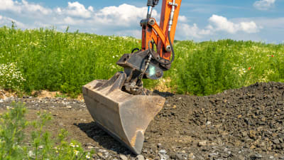 En grävskopa är i beråd att gräva i en fläck brun jord omringad av grön växtlighet på en höjd cirka en halv meter.