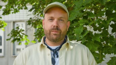 Man i ljusa kläder står framför ett träd