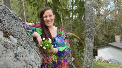 Laura Leppänen, en dam med färggrann klänning och en bukett vitsippor i handen, lutar sig mot en sten.