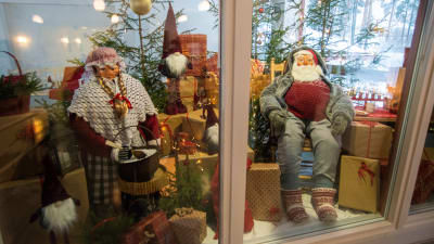 Två stora dockor föreställande julgubben och julgumman sitter omgivna av julklappar.