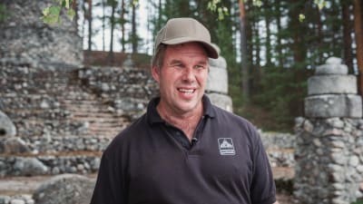Man i skärmmössa och svart t-shirt framför minnesmärke i sten.