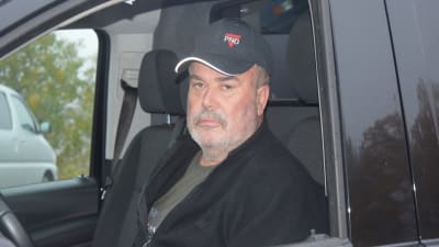 En man med svart keps och jacka sitter bakom ratten på en svart bil.