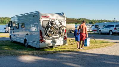 En husbil på en camping. Lättklädda semesterfirare går förbi. 