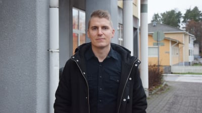 En ung man i kort ljudbrunt hår och svart skjorta med svart ytterkappa på.