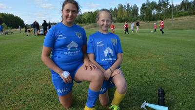 Wilma Mäki och Nea Klingberg, två fotbollsflickor i blå spelskjortor, står på knä vid en fotbollsplan.
