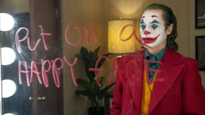 Joker (Joaquin Phoenix) iklädd clownmask står framför en spegel där han skrivit "Put on a happy face" med rött läppstift. 