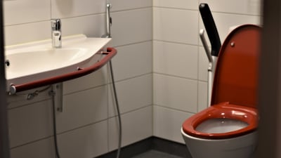En toalett med röd sits och handtag på sidorna. Vita kakel på väggarna och ett vitt handfat med ett rött handtag.