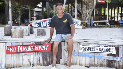 En thailändsk man sitter på en bänk på en strand. Han ler och tittar in i kameran.