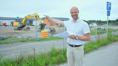 Man i vit kragskjorta framför byggarbetsplats på stor grusplan med stängsel omkring. Mannen pekar mot arbetsplatsen.