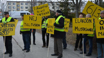 Nio personer står med gula plakat med sloganer mot en kommunsammanslagning.