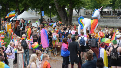 Färggrant klädda personer, regnbågsflaggor och en stor vit uppblåst enhörningsballong på en prideparad.