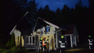 Några brandmän står vid ett förkolnat, ljusgult trähus som förstörts i en brand. 