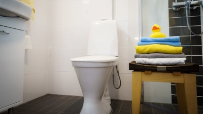 En wc-stol i ett kaklat badrum. Bredvid en pall med vikta handdukar och överst en gul badanka.