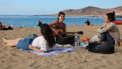 Hiekkaranta, jolla kolme tyttöä istumassa. Keskimmäinen tyttö soittaa kitaraa.