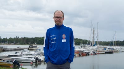 Tränaren för FC Lovisa Niclas Mattson står vid en hamn med båtar i bakgrunden. 