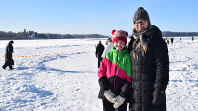 En dotter och hennes mor står vid en sjö som är isklädd. De ler och har på sig vinterkläder.
