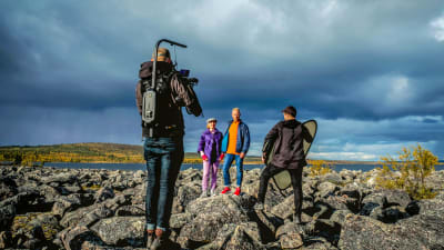 Två personer poserar för tv-kamera på stenar framför en sjö, i förgrunden en kameraman och en hjälpreda.