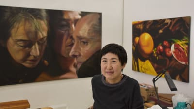 En kvinna sitter i sin atelje omgiven av tavlor som föreställer människor och frukter.