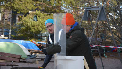 Två män, den ena med turkos mössa och den andra med orange mössa, förbereder sig i en övning att skjuta iväg en nödraket. De skyddar sig bakom en plexiglasskiva.