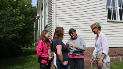 Tre kvinnor och en ung flicka står och talar framför en vit skolbyggnad. De ser glada ut och gestikulerar. 