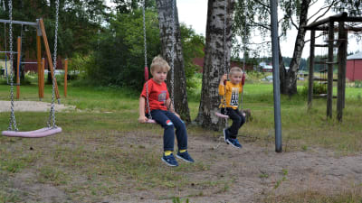 Två pojkar sitter och gungar på en skolgård. I bakgrunden syns träd och en klätterställning.