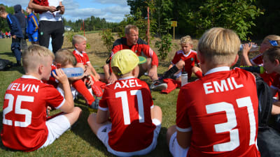 Pojkar i röda fotbollsskjortor sitter runt sin tränare efter en fotbollsmatch. På ryggarna står namnen Melvin, Axel och Emil.