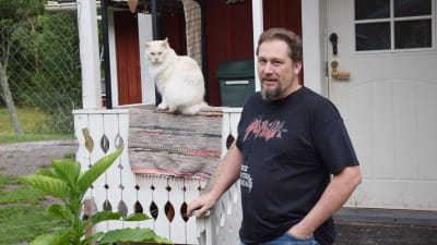 En man står på en trappa i svart t-skorta. Han heter Johan Holmberg. Bredvid honom sitter en vit katt på ett staket. 