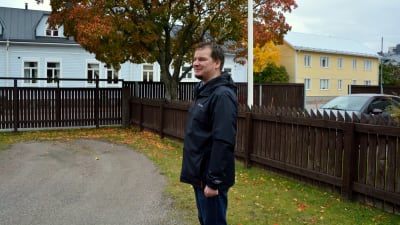 En man står framför ett brunt staket klädd i svart jacka.