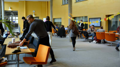 många studenter befinner sig i aulan av asa-huset. där finns stolar och bord i gulaktiga färger