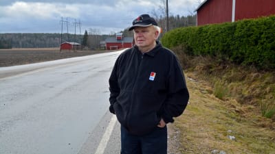 En man i svarta kläder står bredvid en landsväg och en granhäck. 