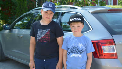 Två pojkar står framför sin gråa bil.